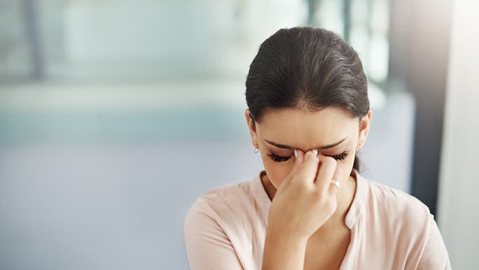 A young woman hangs her head low and pinches between her eyes with two fingers as she ponders the connection between cortisol and stress levels.
