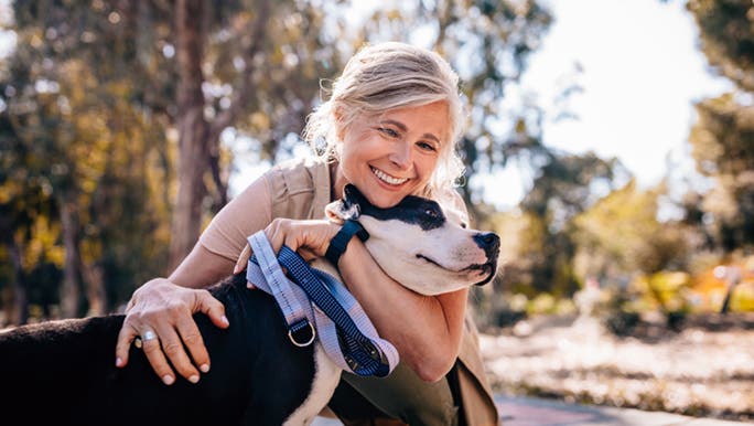 A woman cuddles her dog in on a sunny day. She has grey hair and a big smile on her face. 