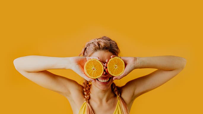 Standing in front of an orange wall, a lady with orange hair holds up two halves of an orange in front of her eyes. 