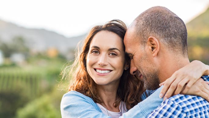 A woman smiles into the camera, she is dating after a divorce and has her arms around her companions shoulders. 