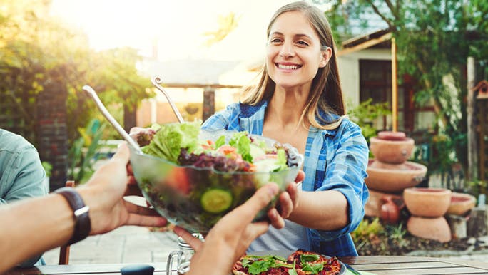 A young woman wearing a blue shirt is smiling as she holds a salad, which is considered good for gut health
