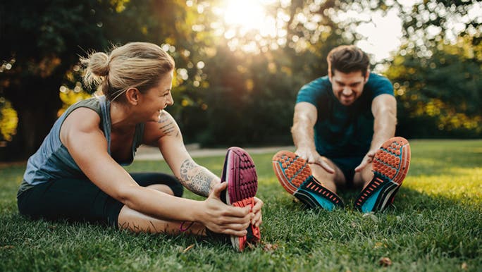 A white Caucasian couple sitting on the grass in a park wearing exercise clothes and stretching, which is a recommended form of exercise when trying to conceive