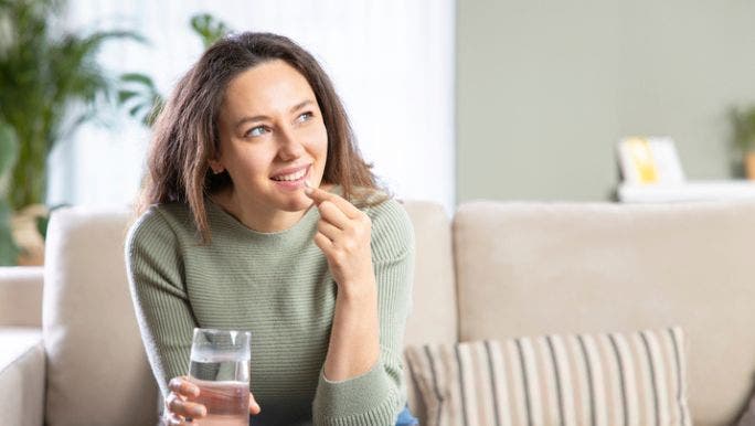 A young woman is smiling on a sofa and holding a glass of water as she takes a probiotic supplement