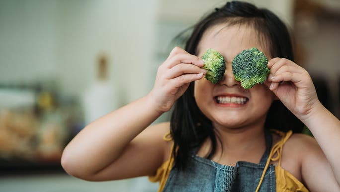Happy little girl holding broccoli florets over her eyes