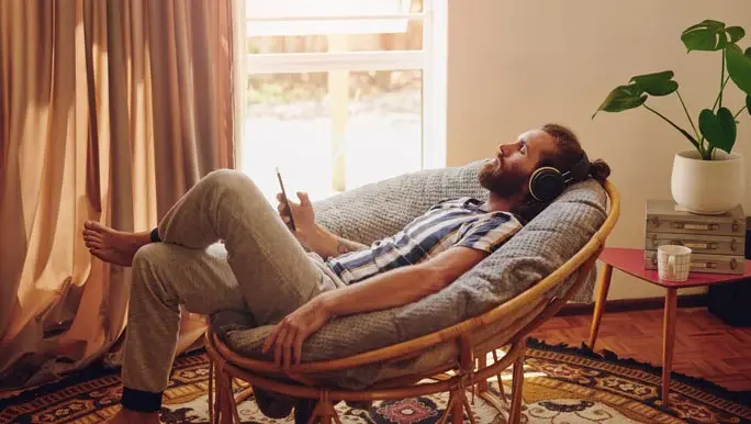 Man relaxing in a cane armchair eyes closed, wearing headphones and listening to music