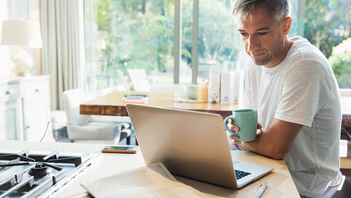 A man with grey hair is working at a laptop and drinking coffee