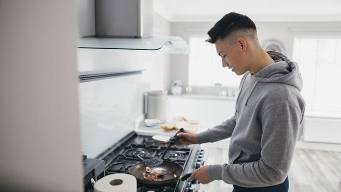 A boy is cooking something on a stove top, it looks to be a protein rich food for teenagers. 