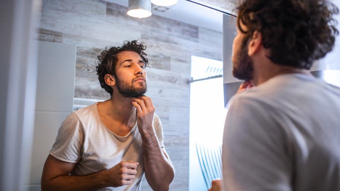 A young man is inspecting his skin in a bathroom mirror