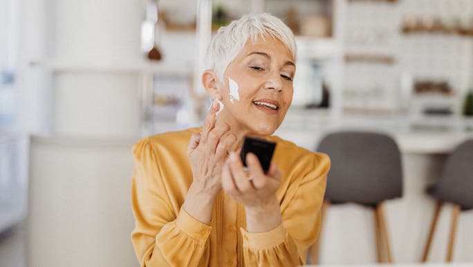 A middle-aged woman with short grey hair and wearing a yellow blouse is applying face cream while looking in a handheld mirror