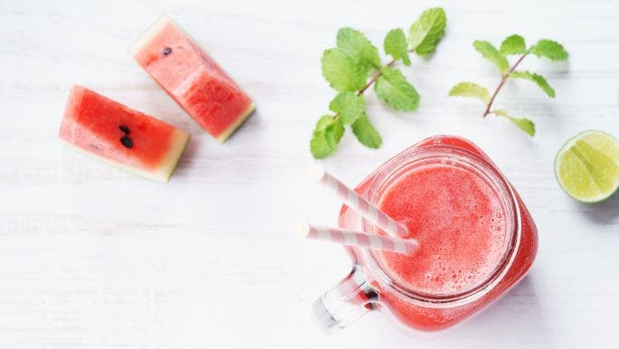 A glass of watermelon and mind juice containing two straws is on a table with cut slices of watermelon and sprigs of mint nearby