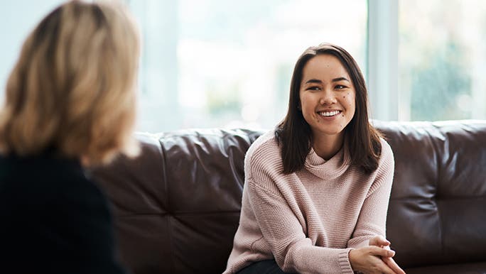 A woman is sitting on a couch talking to another woman. You can only see the blurred back of the second woman, they seem to be having some sort of health check up.