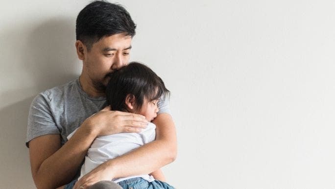 An Asian father is sitting and holding his young child who is asleep
