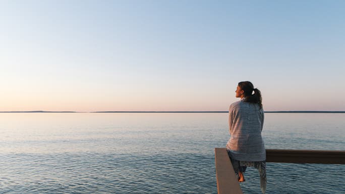 A lady perches on a handrail in front of the ocean. She is staring at the sunset looking for her motivation. 
