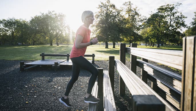 An older woman is doing step ups in the park on a bench which are one of the best exercises for your glutes. 