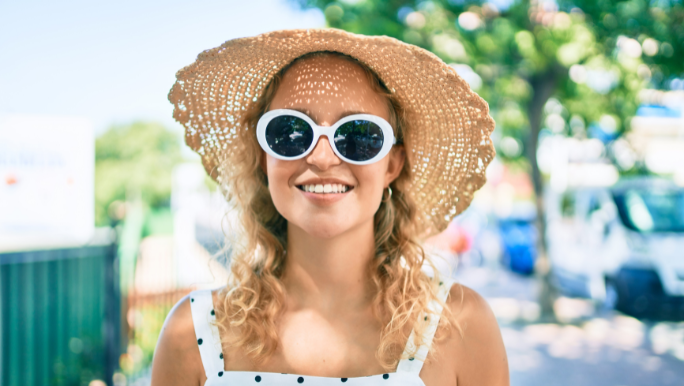 A young woman is wearing a hat and sunglasses and smiling as she stands outdoors on a sunny day