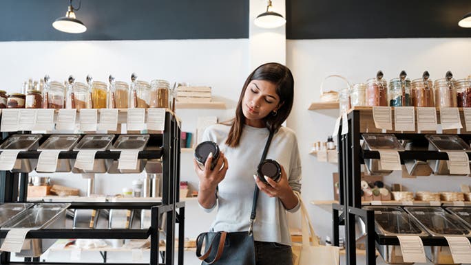 A woman is in an eco healthy food store, holding up two glass jars that are filled with produce. She’s trying to decide which one to buy.