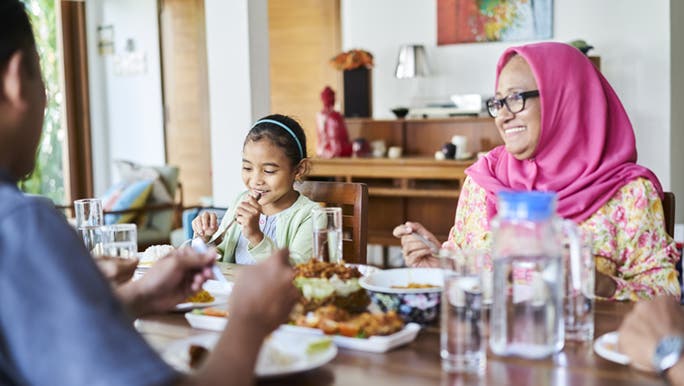 A family sits down to dinner, the woman is wearing a pink scarf around her hair and the little girl is smiling and eating happily. 