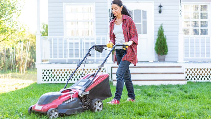 A lady in a red shirt is mowing the lawn in front of a weatherboard house. She is enjoying the incidental activity.