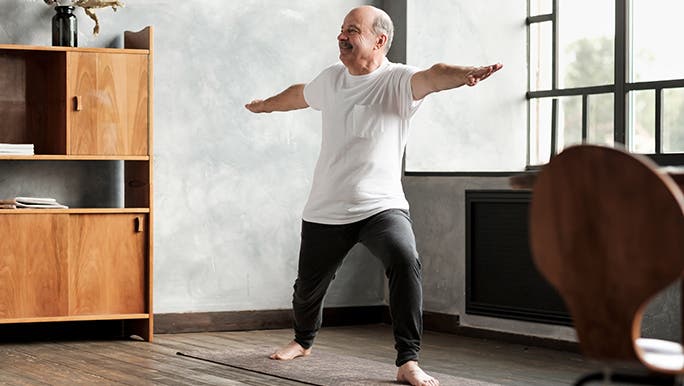 A man does Yoga in his home, he is wearing track pants, a t-shirt and no shoes. 