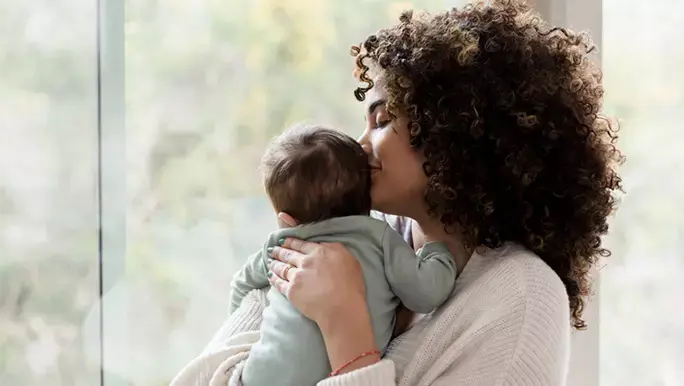 Young mom gives her baby a kiss on his head while standing next to a window in their home