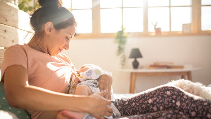A woman with dark hair and a pink top relaxing on a bed while breastfeeding her baby