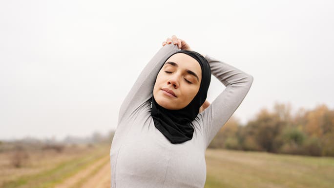 A woman is standing on a dirt track, stretching with her eyes closed. It looks like she is going for a run. 