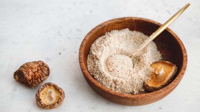 A  flat lay image of mushroom powder in a wooden bowl surrounded by shiitake mushrooms