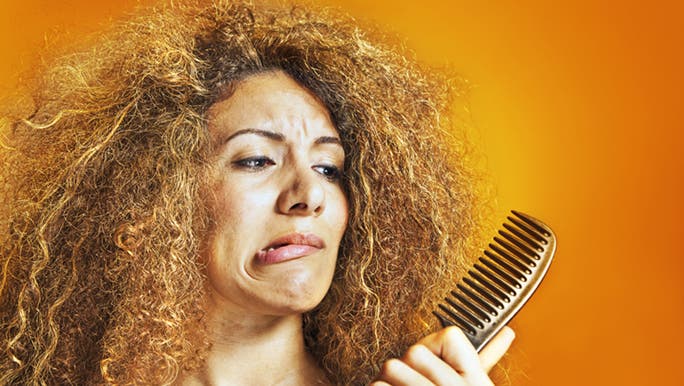 A woman stands in front of an orange wall, trying to comb her messy hair, wondering how to avoid frizzy hair when sleeping.