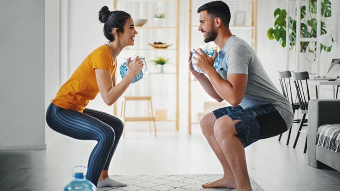 While doing squats, one of the best compound exercises, a couple are smiling and laughing at each other during their workout in the lounge room.