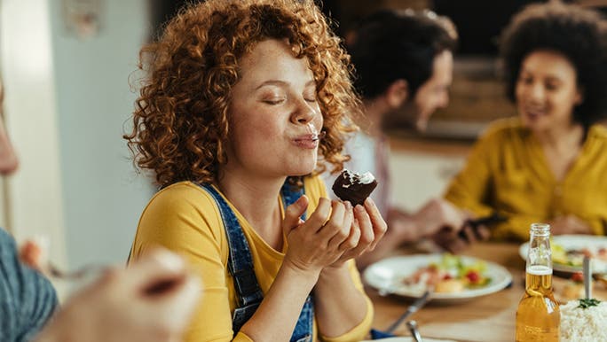 A woman with red curly hair and wearing a yellow top is closing her eyes as she chews her food, eating mindfully