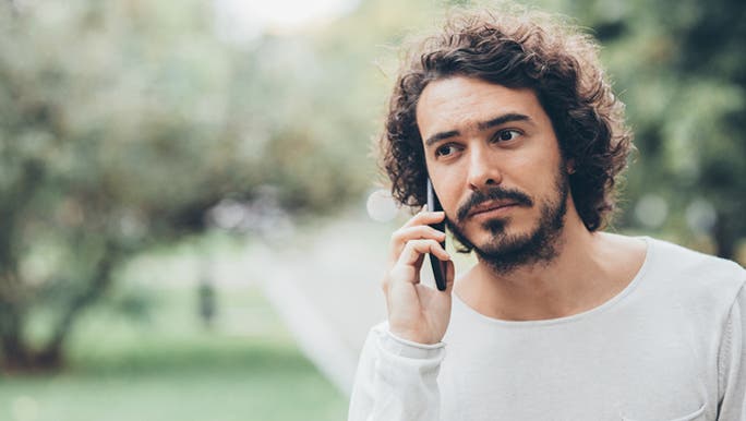 A dark-haired young man with facial hair wearing a white top is standing in a park having a phone conversation and looking sad