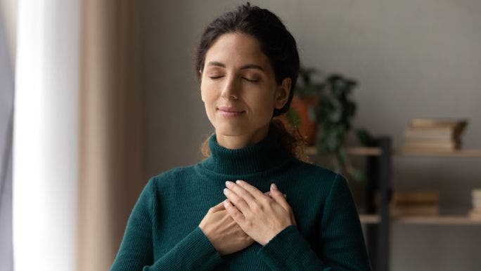 A young woman with eyes closed and her hands placed across her chest is smiling and practising words of affirmation