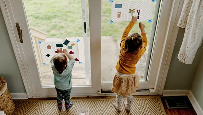 Two children do an art project. There is paper stuck to a glass door and they are putting stickers on them. 