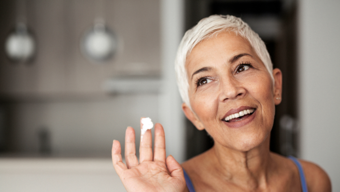 A senior woman is smiling and holding a small amount of face cream on her finger