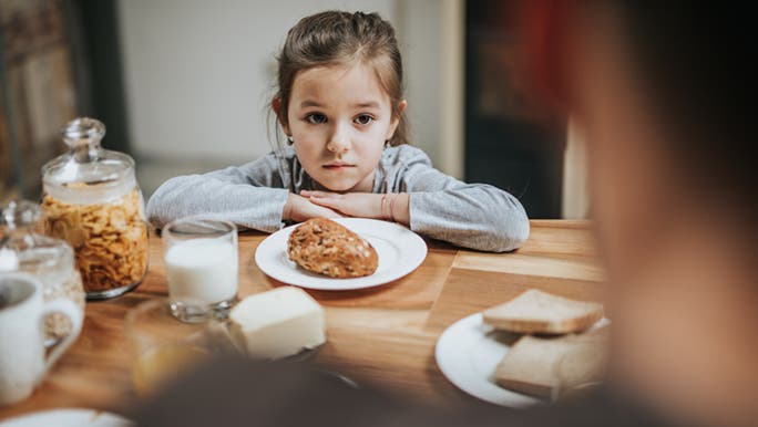 A child who is a fussy eater sits, unenthusiastically at a kitchen table. There is a wholemeal bread roll on her plate and it doesn’t look like she will be eating it.