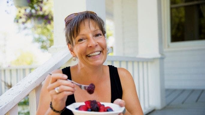 A Caucasian woman is smiling as she eats a bowl of blueberries and strawberries