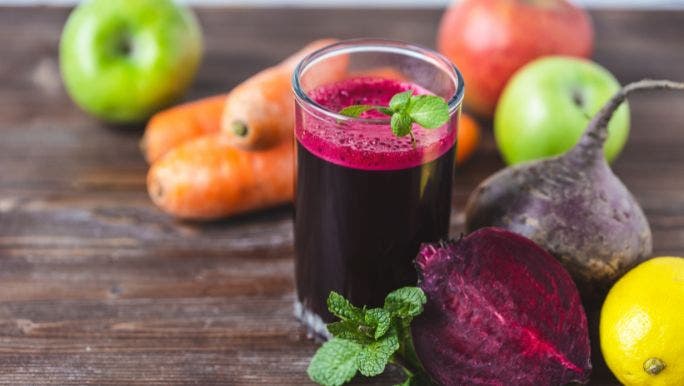 A glass of fresh juice sits on a wooden table surrounded by apples, carrots and beetroot