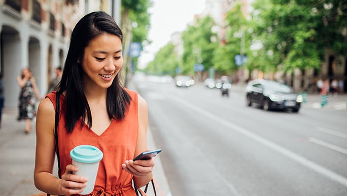 A woman smiles and looks at her phone, she has a coffee in one hand and looks to be taking a work-life balance break. 
