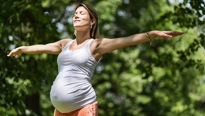 A pregnant woman stands with her arms outstretched in front of a huge leafy tree. Her eyes are closed and it looks like she may be doing some yoga pelvic floor exercises. 