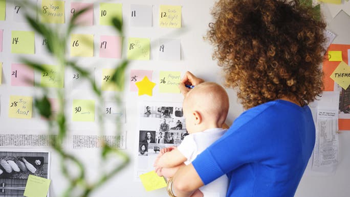 From behind, a woman holds a baby while writing on colourful Post-it notes that are stuck to the wall. 