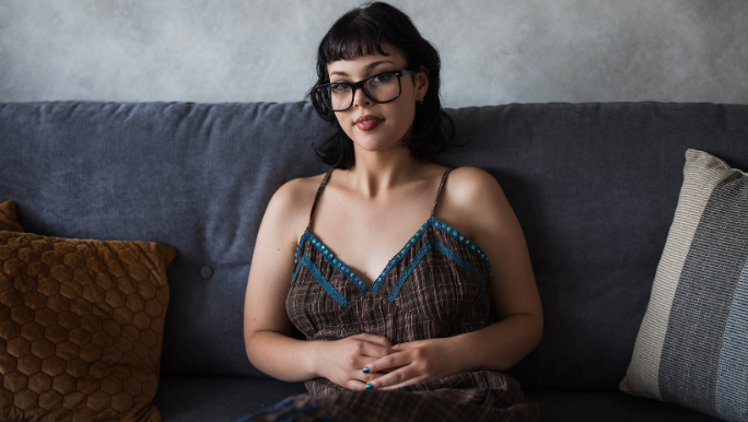 A young woman wearing black glasses is sitting on a sofa with her hands on her tummy