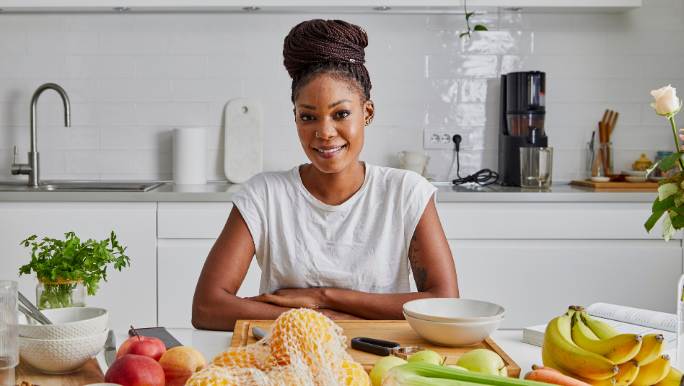 A young black woman is standing at a kitchen bench filled with fresh fruit and smiling