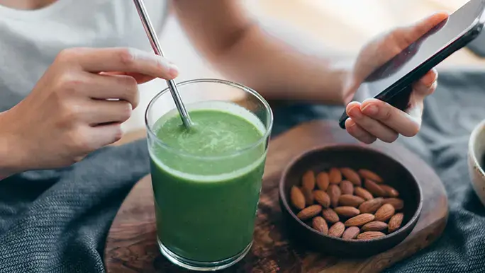 Woman (head cropped out) sits down at table while holding her phone in one hand ad holding her metal straw in the other. The straw is in a cup holding green powder mixed with water. Next to it is a dish filled with almonds.