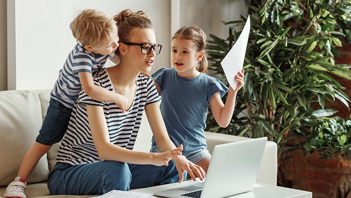 A mother works on a laptop as her two children climb all over her, she looks stressed. 