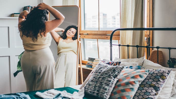 A woman with long dark hair is looking at herself in a full-length bedroom mirror as she fixes her hair