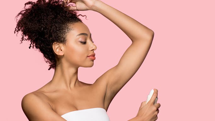 In front of a pink background, a wonder wearing a towel around her chest lifts her arm to apply the best natural deodorant in Australia.