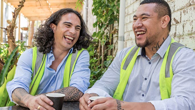 Two dark-skinned men wearing hi-vis vests sharing a cup of coffee and smiling as they chat while having a break from work