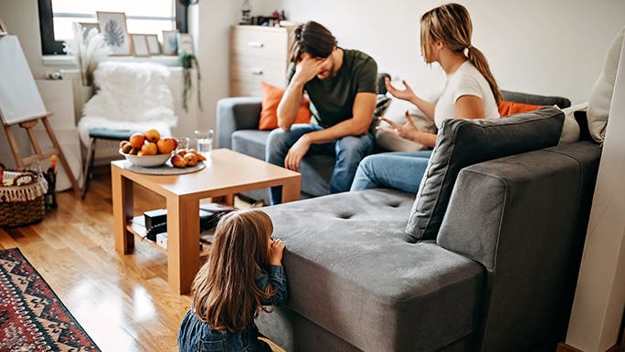 A couple argues on a couch as a child sits on the ground watching. There is a lot of relationship conflict in the picture. 