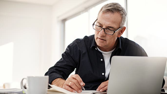 A man in glasses is concentrating on some notes he is taking. There is an open laptop in front of him, he looks deep in thought. 
