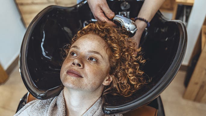 A woman with red hair  is smiling while she has her hair washed at a hairdresser, happy she knows how to stop frizzy hair after washing.